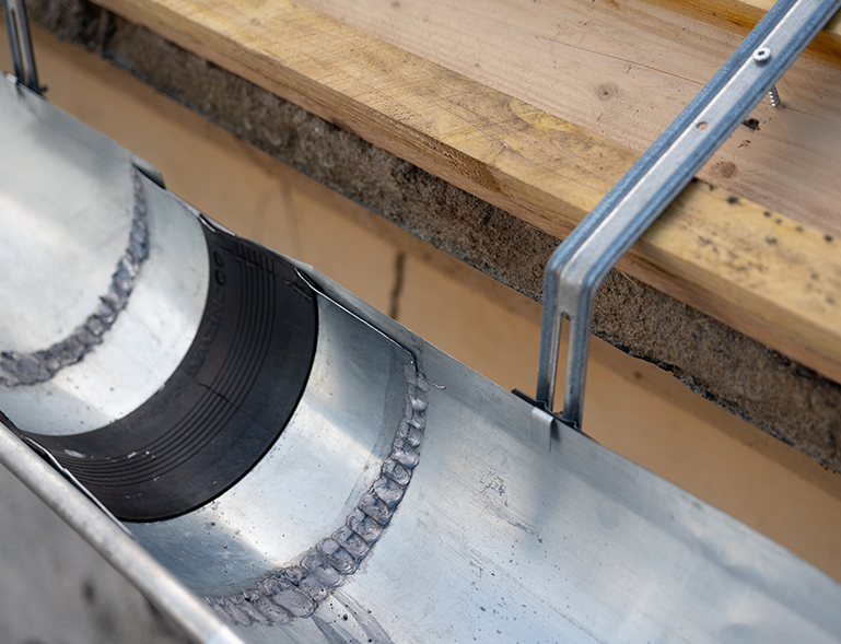 Gros plan d’une gouttière en acier galvanisé montrant une soudure entre sections, un insert noir d’étanchéité et des supports métalliques vissés sur la planche de rive en bois.